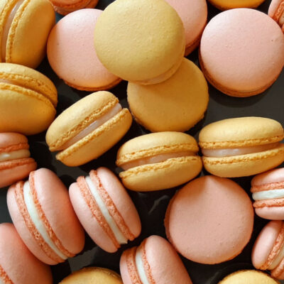 Auf dem Bild sind mehrere Macarons in den Farben Gelb und Rosa mit einer hellen Füllung zu sehen. Die Macrons sind ungeordnet auf einer schwarzen Platte angerichtet.