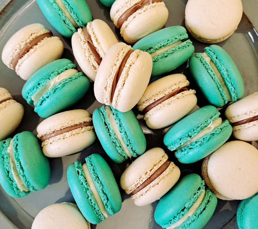 Auf dem Bild sind mehrere Macarons in den Farben Hellbraun und Türkis mit einer hellen und dunklen Schokoladenfüllung zu sehen. Die Macrons sind ungeordnet auf einer grauen Platte angerichtet.