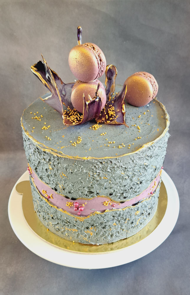 Auf dem Bild ist eine Cremetorte im Design eines Faultline Cakes, das heißt in der Mitte der Torte befindet sich zwei getrennte Cremekanten in den Farben grün und weiß zu sehen. Die Torte ist an der Seite mit verschieden großen Zuckerperlen in den Farben Gold, Weiß, Bordeaux und grün dekoriert. Auf der Oberfläche der Torte befindet sich ein filigranes Zuckerstück , welches mit 3 Macarons bestückt ist. Das Zuckerstück weist einen leichten marmorierten Farbton in Dunkel- und Hellgrün auf und ist mit einem Goldenen Rand versehen.