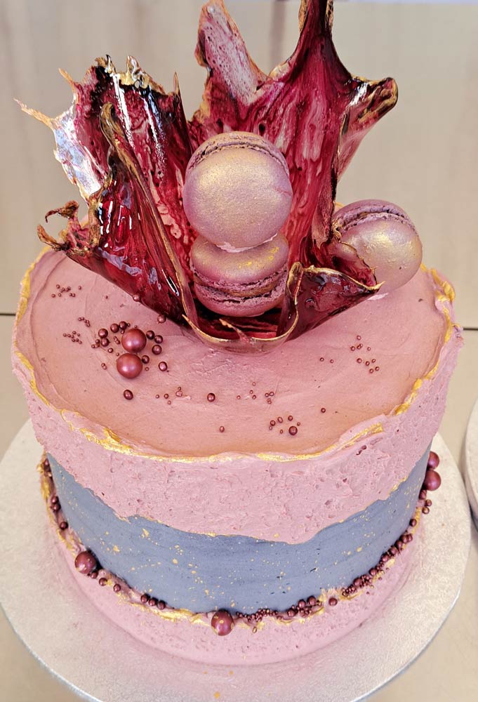 Auf dem Bild ist Cremetorte im Design eines Faultline Cakes, das heißt in der Mitte der Torte ist eine Bruchline aus Buttercreme in der Farbe grau zu sehen . Die Torte ist hauptsächlich in der Farbe Rose und grau gestaltete und an der Seite mit Zuckerperlen in der Farbe Bordeaux Metallic dekoriert. Auf der Oberfläche der Torte befindet sich ein filigranes Zuckerstück , welches mit 3 rosa Macarons bestückt ist. Das Zuckerstück weist einen leichten marmorierten Farbton in Dunkel- und Hellbordeaux auf und ist mit einem Goldenen Rand versehen.