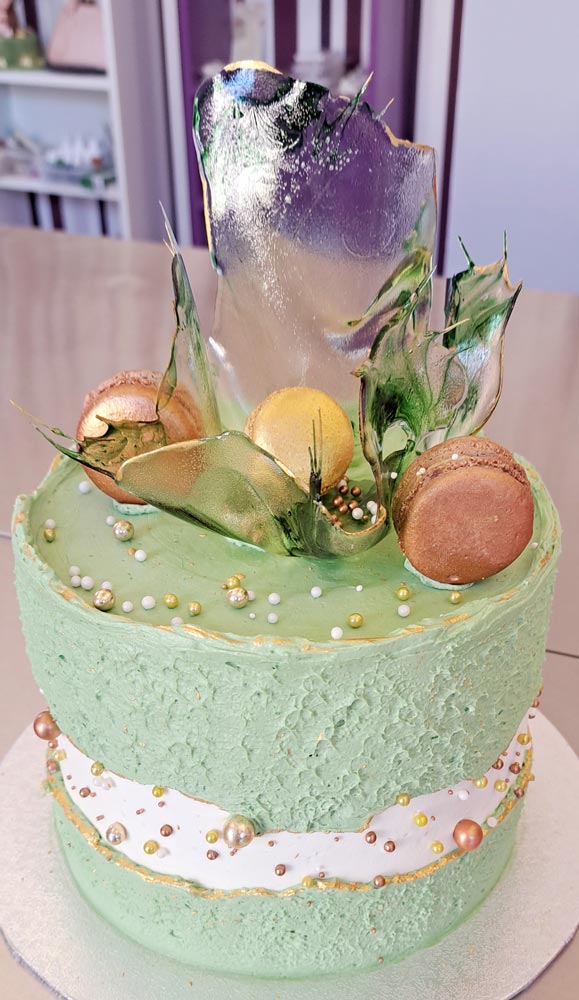 Auf dem Bild ist ein Ausschnitt einer Cremetorte im Design eines Faultline Cakes, das heißt in der Mitte der Torte befindet sich zwei getrennte Cremekanten in den Farben grün und weiß, zu sehen. Die Torte ist an der Seite mit verschieden großen Zuckerperlen in den Farben Gold, Weiß, Bordeaux und grün dekoriert. Auf der Oberfläche der Torte befindet sich ein filigranes Zuckerstück , welches mit 3 Macarons bestückt ist. Das Zuckerstück weist einen leichten marmorierten Farbton in Dunkel- und Hellgrün auf und ist mit einem Goldenen Rand versehen.
