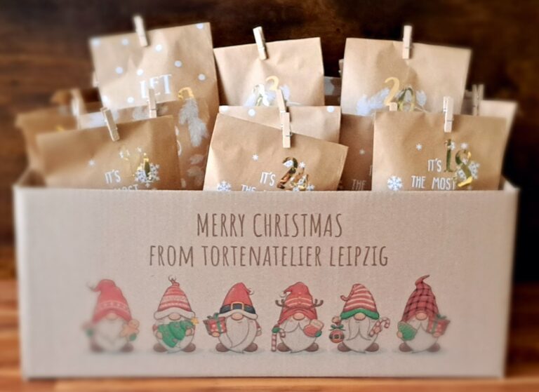 Rechteckiger brauner Karton mit dem Schriftzug Merry Christmas from Tortenatelier Leipzig und Aufdruck von 6 Weihnachtswichtel unter dem Schriftzug. Der Karton ist mit weihnachtlichen Adventstüten, welche mit einer Holzklammer geschlossen wurden, gefüllt.