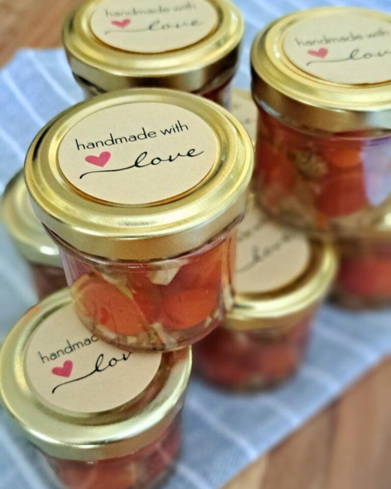 Zu sehen sind mehrere kleine Gläser gefüllt mit Tomaten, Zwiebeln und anderen Zutaten. Die Gläser stehen auf einen blau-grauen Tuch mit weißen Streifen .Auf dem goldenen Glasdeckel ist der Schriftzug Handmade with love zu sehen