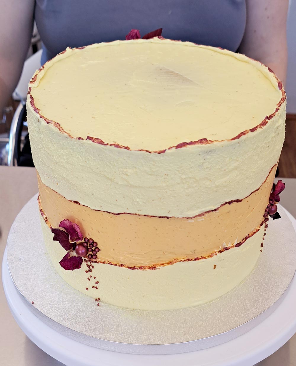 Auf dem Bild ist Cremetorte im Design eines Faultline Cakes, das heißt in der Mitte der Torte ist eine Bruchline aus Buttercreme in der Farbe orange zu sehen . Die Torte ist hauptsächlich in der Farbe Pastellgelb , Orange und Bordeaux gestaltet. Die Bruchlinie ist in der Farbe Bordeaux gestaltet , genauso wie die Seitendekoration aus Zuckerperlen und Blütenblättern in der gleichen Farbe gestaltet ist.