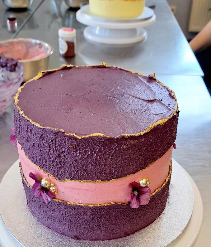 Auf dem Bild ist Cremetorte im Design eines Faultline Cakes, das heißt in der Mitte der Torte ist eine Bruchline aus Buttercreme in der Farbe Rosa zu sehen . Die Torte ist hauptsächlich in den Farben Lila , Rosa und Gold gestaltet. Die Bruchlinie ist mit Gold abgesetzt . Die Seitendekoration besteht aus goldenen Zuckerperlen und essbaren Blüten. Es sind rosafarbene Hornveilchen.
