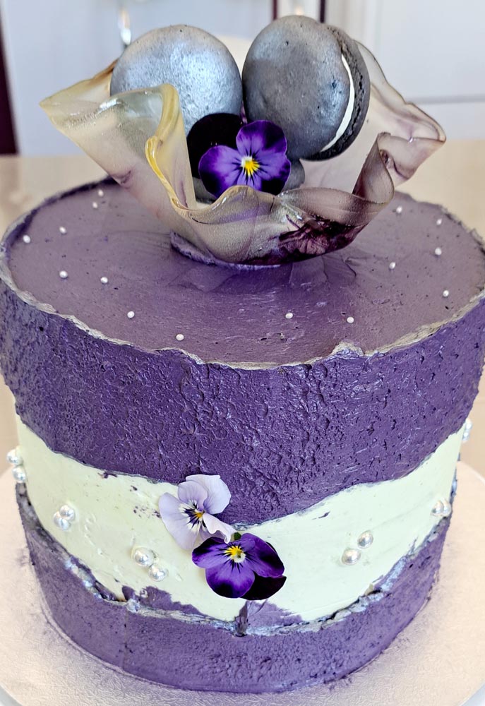 Auf dem Bild ist Cremetorte im Design eines Faultline Cakes, das heißt in der Mitte der Torte ist eine Bruchline aus Buttercreme in der Farbe mintgrün zu sehen . Die Torte ist hauptsächlich in der Farbe Lila und grau gestaltet und an der Seite mit silbernen Zuckerperlen und 2 Hornveilchen dekoriert. Auf der Oberfläche der Torte befindet sich ein filigranes Zuckerstück . Es ähnelt einer gewellten Glasschale, welche in der Mitte mit 3 grauen Macarons und einem blauen Hornveilchen bestückt ist. Das Zuckerstück weist einen leichten marmorierten Farbton in Dunkellila und Grau auf .